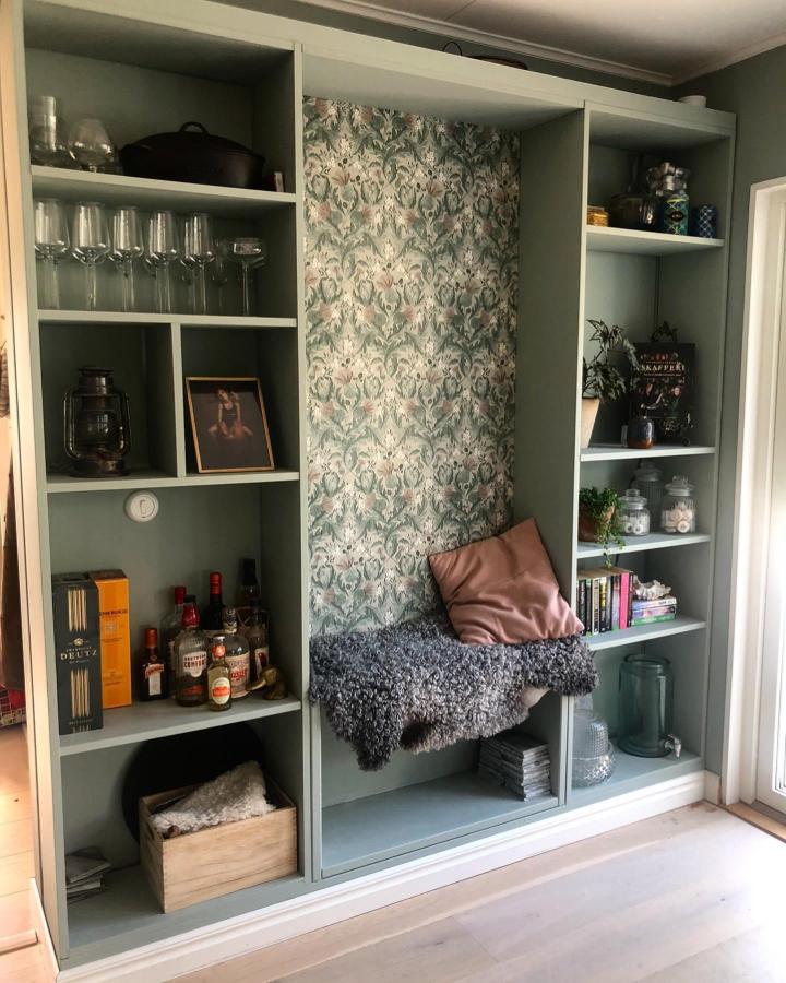 Soft sage green painted bookshelf with a cozy built-in bench and floral wallpaper from @renoveringsfamiljen creating a snug reading corner.