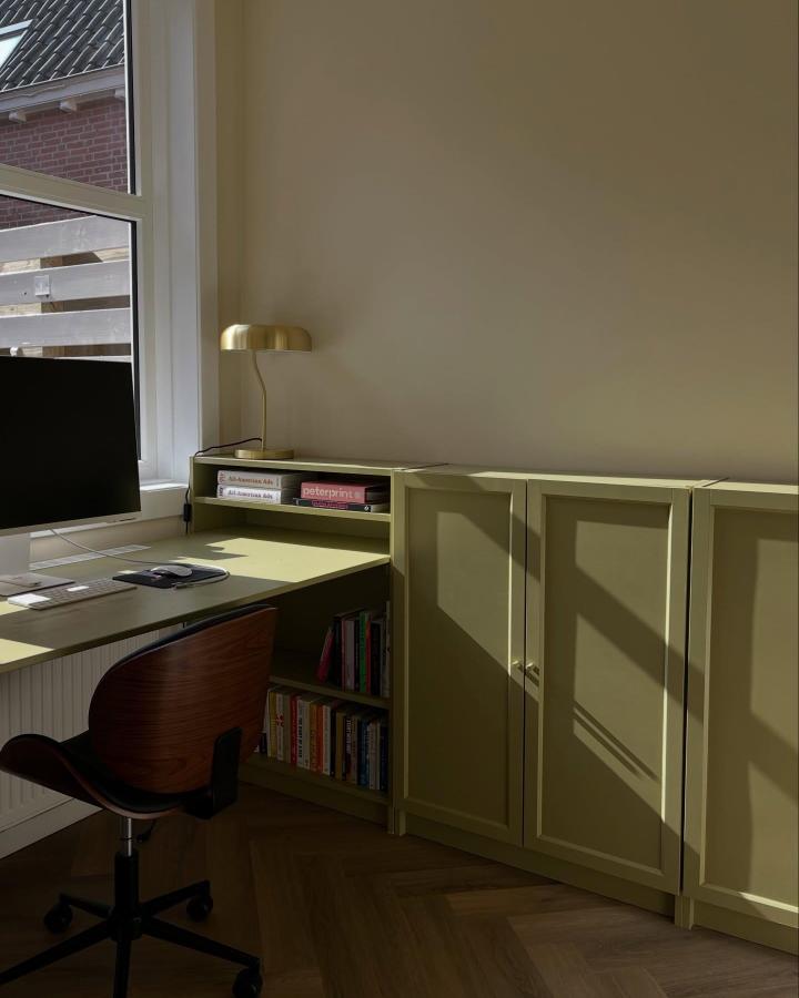 Soft olive green bookcase and desk setup from @home.in.the.city with built-in shelving and brass lamp in natural light.