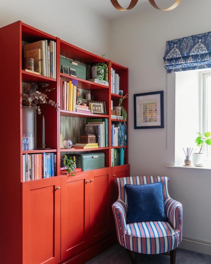 Red painted Billy bookcase with brass knobs and colorful books in cozy reading nook from @rosieandthefarm. Classic style with vibrant, warm finish.