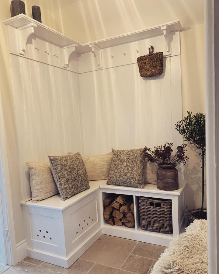 White painted corner bench and storage with hooks in entryway by @ourmedievalcottage. Cozy rustic style with cushions and wicker basket.