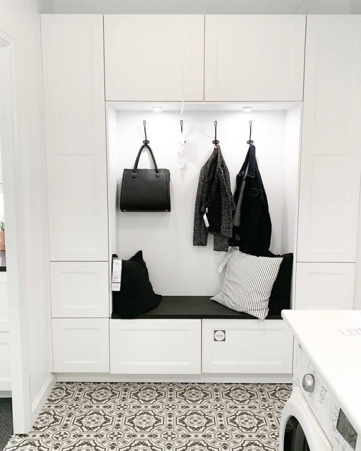 White built-in storage locker seating nook with black cushions and hooks from @ikankitchens. Functional entryway design with overhead cabinets and patterned tile floor.