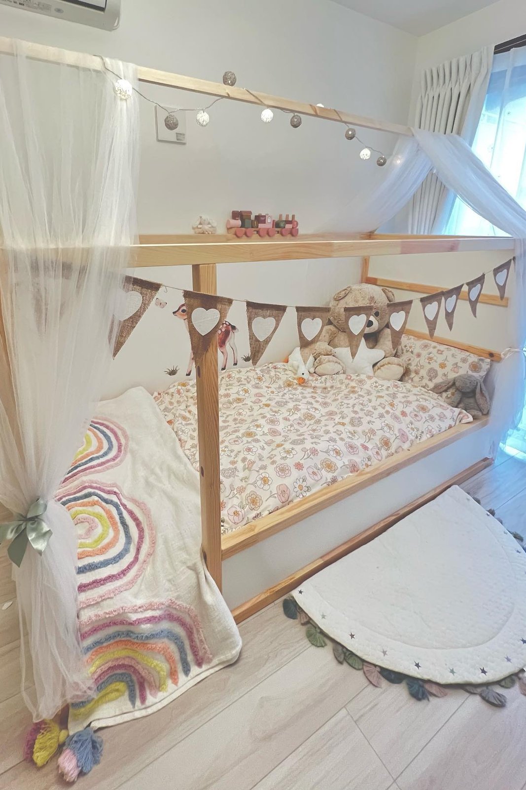 KURA bed with sheer canopy curtains, fairy lights, floral bedding, heart bunting, toys, and a rainbow throw