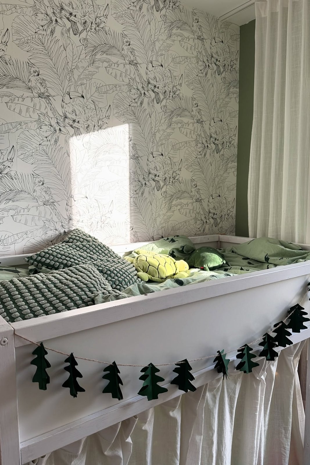White KURA bed with under-bed curtain, tree garland, botanical wallpaper, green bedding, and plush cushions