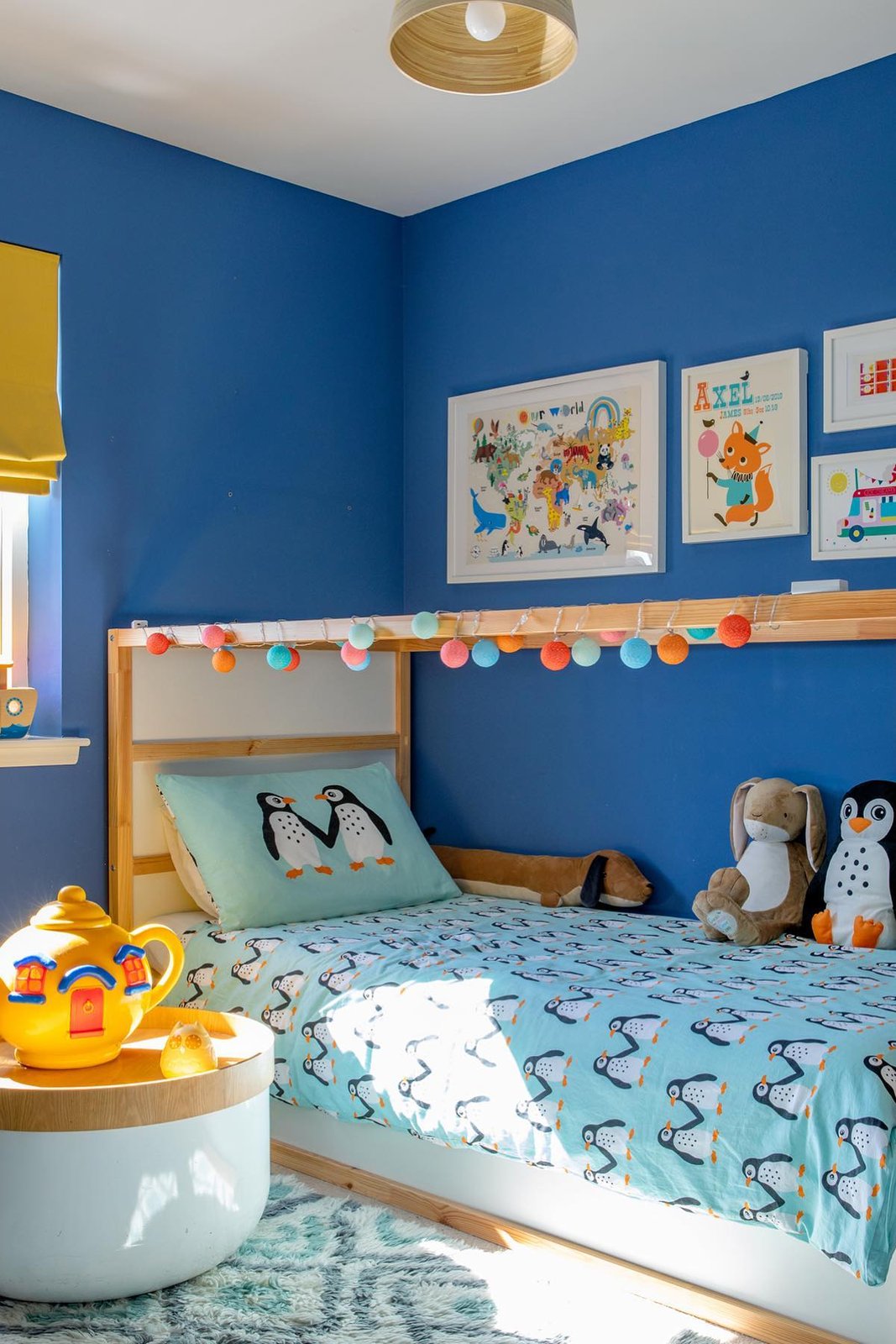 Colorful Pom-pom lights and penguin bedding brighten a natural wood KURA bed in a playful blue child’s room