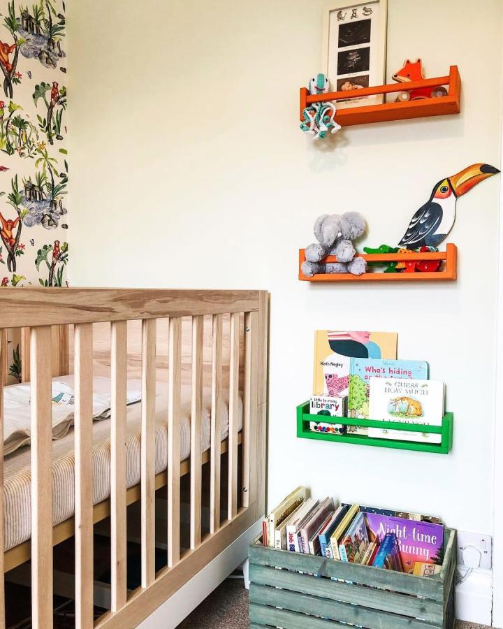 Natural pine and colorful painted wooden racks nursery storage from @hellojenniferhelen with books and toys on white wall next to crib.