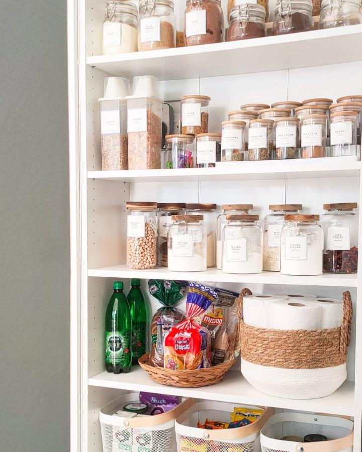 Clear containers with wooden lids on white shelves from @hillo.hajar showing a tidy, natural pantry organization with labels.