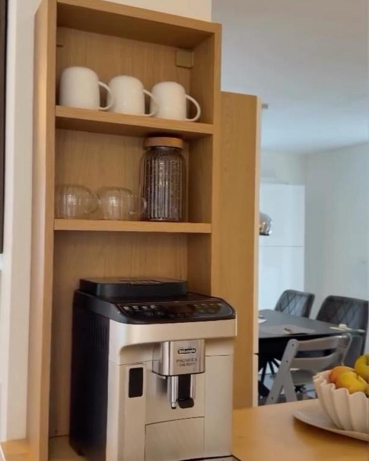 Light natural wood coffee nook with white mugs and glass cups by @laura.kwtr. Streamlined setup with a coffee machine and warm wooden shelves.