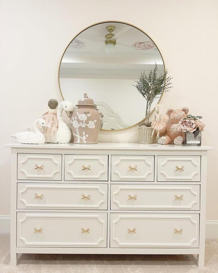 Creamy white IKEA Hemnes dresser with trim overlays and gold bow knobs from @wrapped_in_lace ikea pottery barn dupes in a nursery setting.