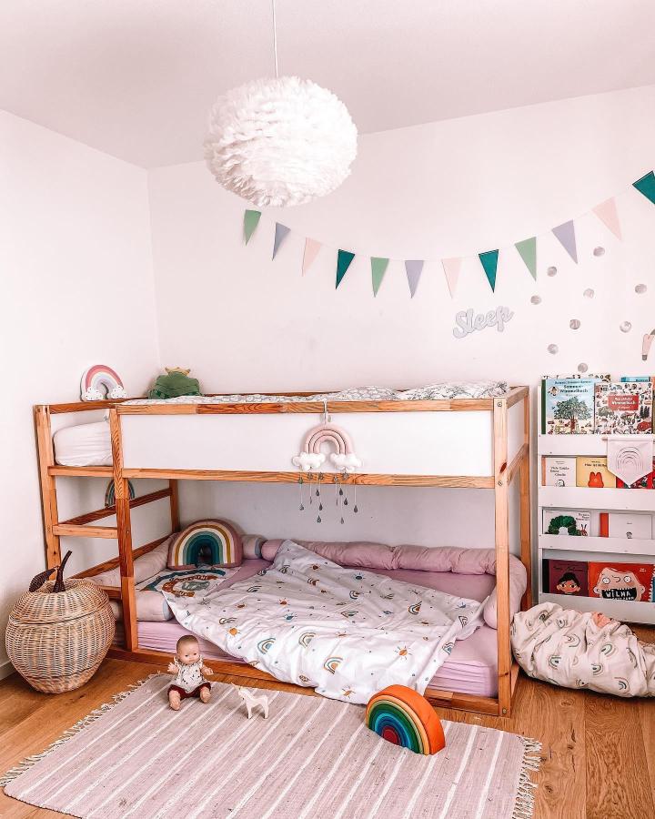 Warm wood rainbow-themed Kura bunk bed from @evaandthegirls. Pastel bunting, rainbow mobile, layered bedding, and reading storage.