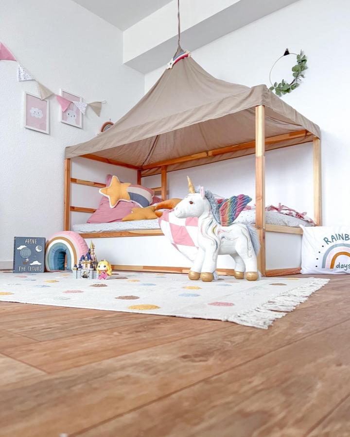 Taupe canopy KURA bed from @kuralove.de. Pink cushions, rainbow pillows, and unicorn toys create a cozy play nook.