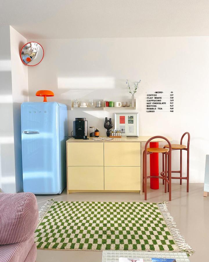 Pastel yellow coffee bar from @studiomysa.nl. Retro nook with blue fridge, red stools, and checkerboard rug.