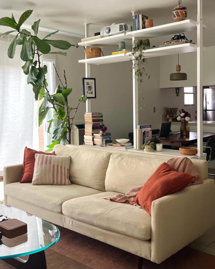 White open-shelf living room divider from apartment therapy. Ceiling-mounted ELVARLI posts separate a sofa and dining area.
