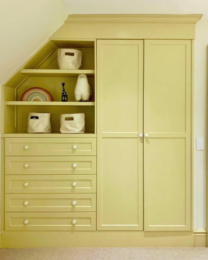 Buttery-yellow built-in nursery wardrobe from @blacksheepid. Sloped ceiling, drawers, shelves, and white knobs.