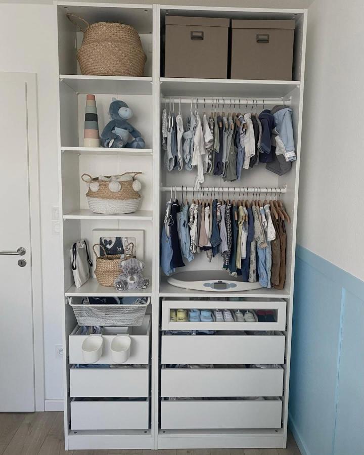 White open-shelving nursery wardrobe from @just.ev. Wicker baskets, taupe bins, and neatly arranged baby clothes.