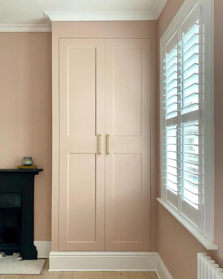 Blush paneled wardrobe from @prettylondonliving. Compact fitted storage beside a bright shuttered window.