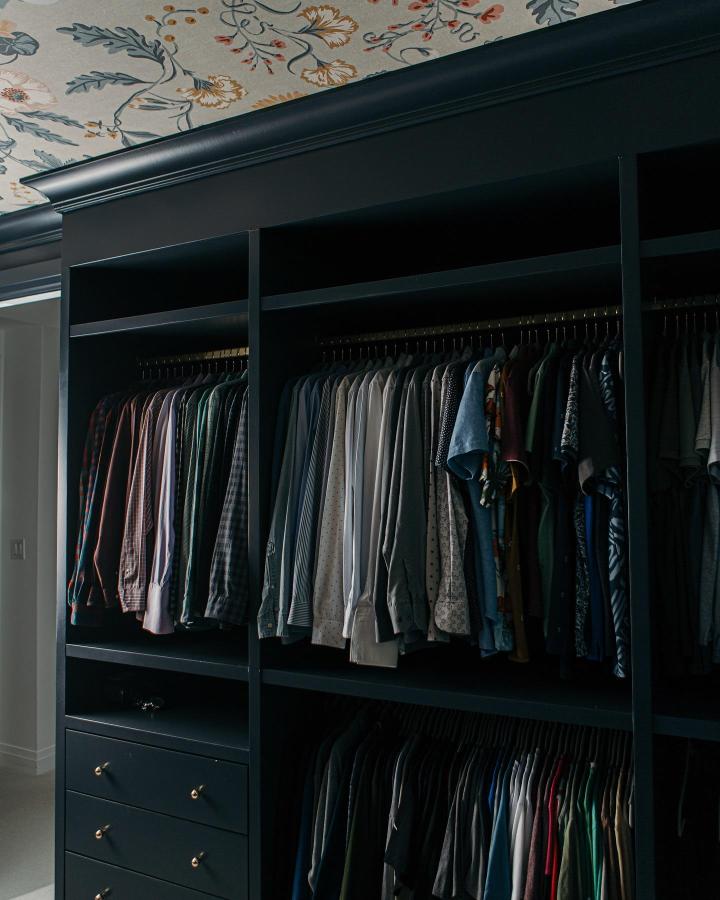 Moody black walk-in wardrobe from @thecumberlandproject. Floral ceiling, double hanging rails, open shelves, and brass drawer pulls.