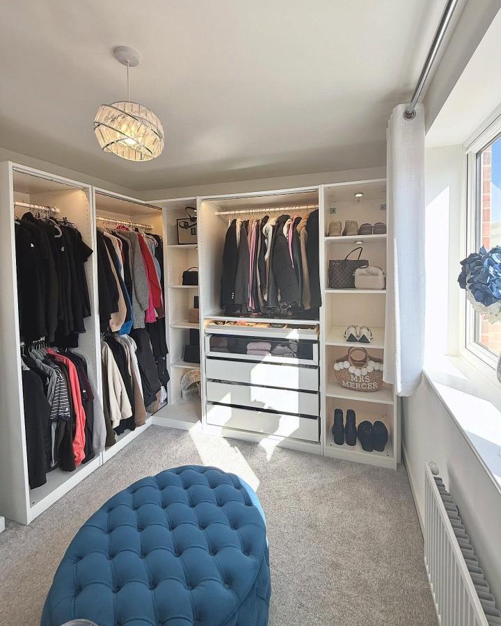 White open PAX walk-in wardrobe from @athomewithkirsty_. Drawers, clothing rails, shoe shelves, and blue ottoman fill a sunny dressing room.