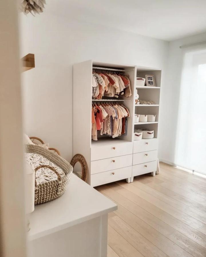 Soft white nursery wardrobe from @eline.vandingenen. Open shelving, baby clothes, drawers, and woven baskets create a calm organized corner.