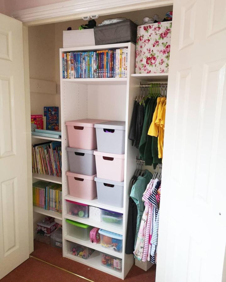 White open shelving wardrobe from @a_mummas_life. Pink storage boxes, books, and colourful children’s clothing organise a Platsa closet.