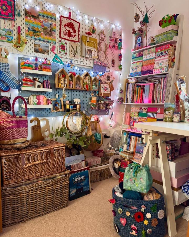 White pegboard craft room from @nanaseamakes. Colorful shelves, yarn, baskets, and fairy lights fill the compact workspace.