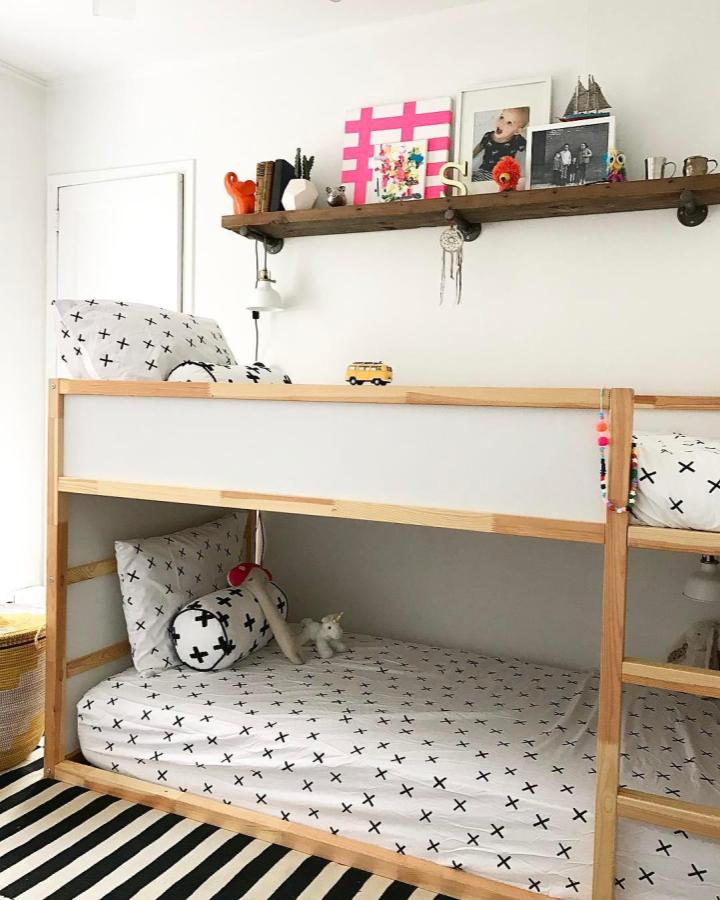 Natural wood bunk bed from @thayerwoodshome. Shared childrenโs room with cross-print bedding, shelf display, and striped rug.