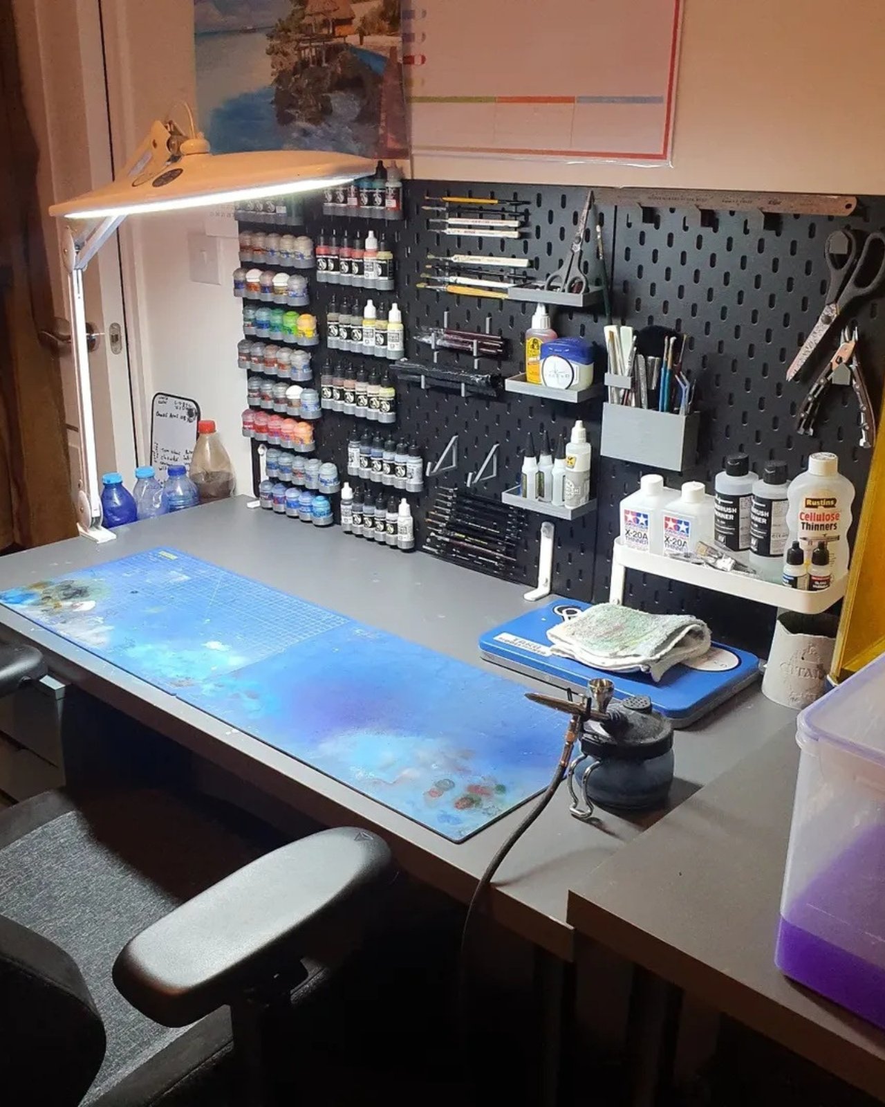 Black pegboard craft desk with paint bottles, brushes, scissors, and organized art supplies under a task lamp