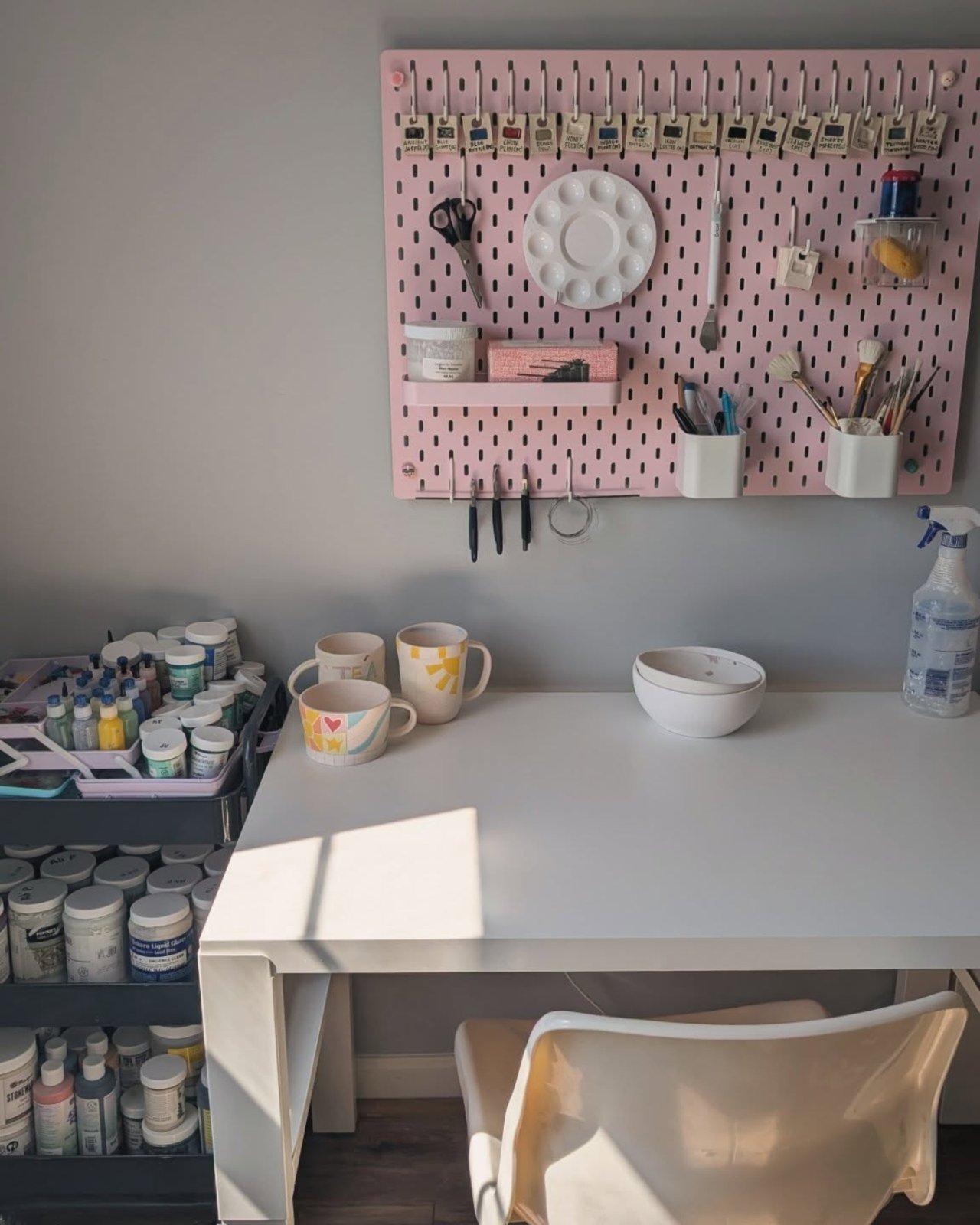 Pink pegboard craft organizer with labeled paint tubes, brushes, palette, cups, and supplies above a white desk